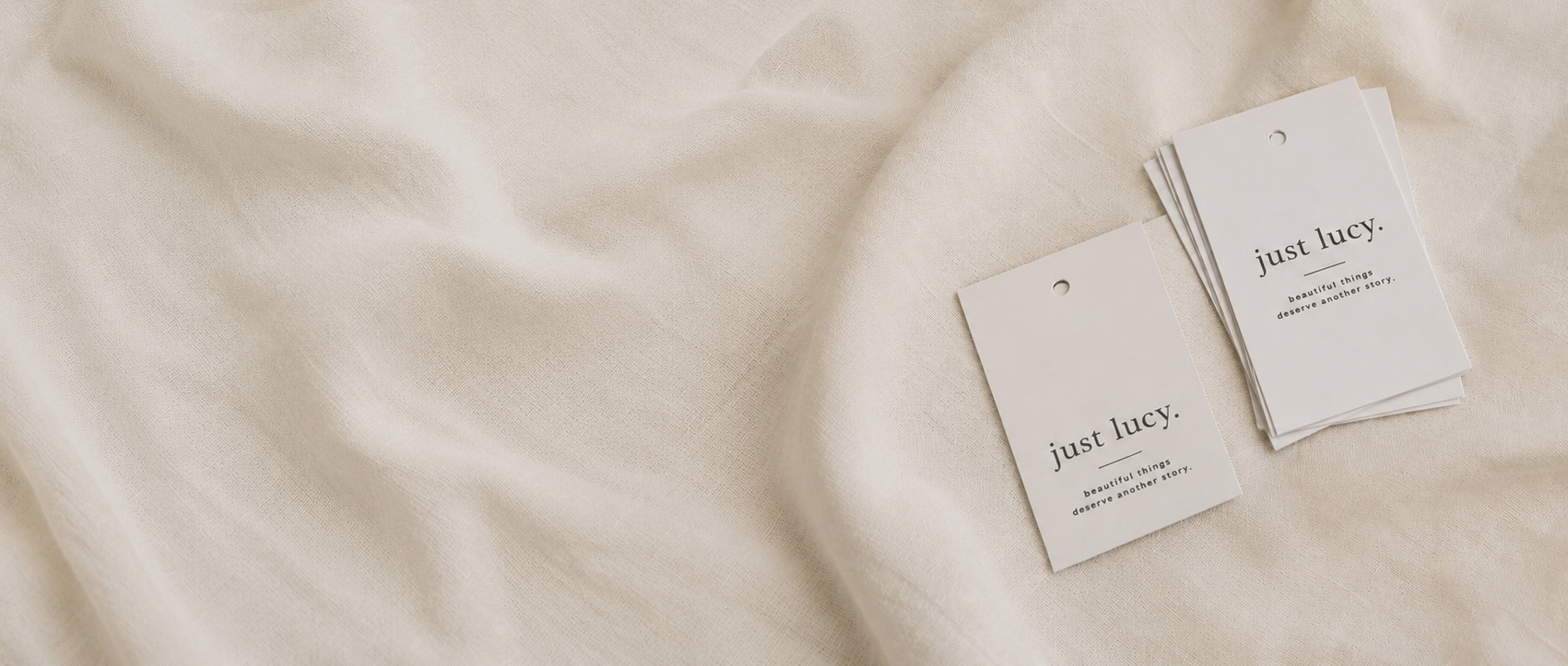 Two small white paper cards printed with the brand name 'just lucy.' and fine text, placed on soft draped cream fabric.